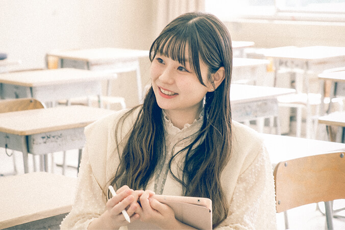 ロングヘアの女性ライターが教室内で椅子に座っています。ノートを手に笑顔で写っている写真です。