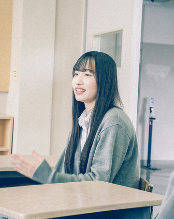 教室内の椅子に座り、笑顔でインタビューを受けているBさんの写真です