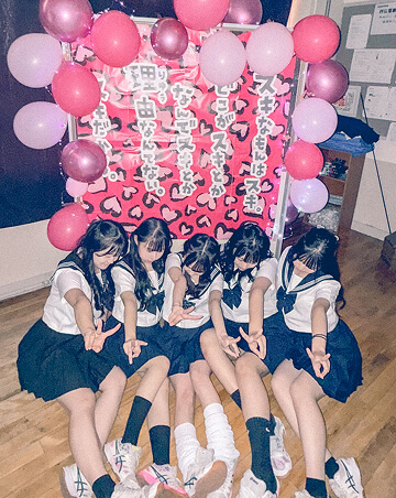 文化祭の写真です。ピンクの風船で飾り付けられたフォトスポットの前に、セーラ服を着た生徒五人が座ってピースをしています。顔は見えないように全員俯いています